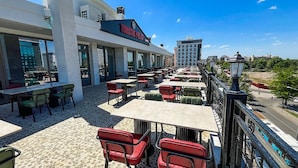 An outdoor seating area with tables and chairs, a building with a sign reading "Turistik Palas," and a view of the street and buildings in the background. - TURİSTİK PALAS OTEL (Diyarbakir)