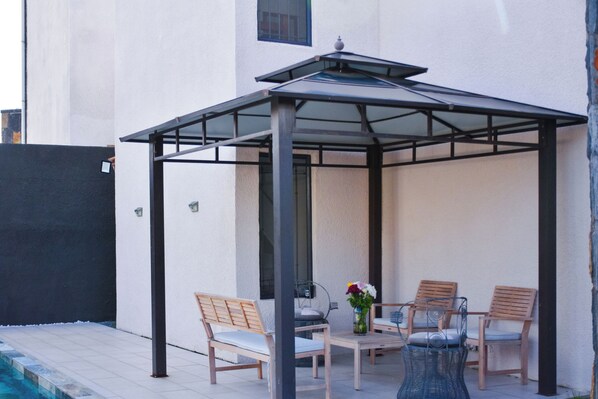 A covered patio area with a table and four chairs, a vase with flowers, and a pool in the background.