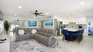 A modern living room with a sectional sofa, a dining area with blue chairs, and a kitchen with white cabinets and stainless steel appliances. - Island 3 bed 3 bath heated pool golf cart kayaks (Indian Rocks Beach)