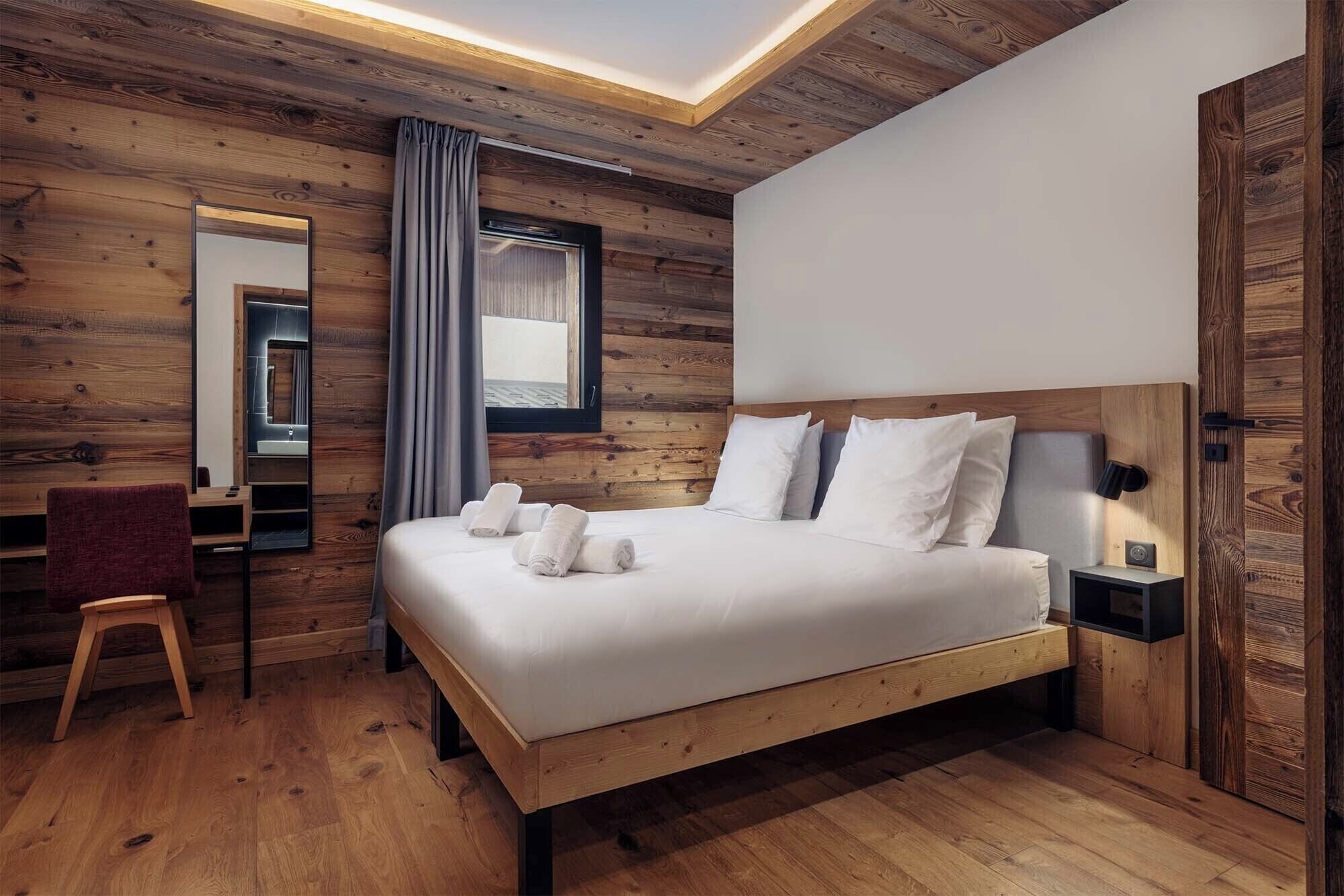 A bedroom with a wooden ceiling, a bed with white linens, a bedside table, a chair, and a mirror.