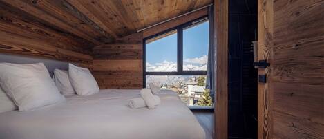 A wooden cabin bedroom with a bed, pillows, and a window overlooking a snowy mountain.