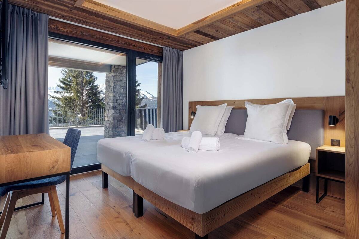 Ein modernes Hotelzimmer mit einem großen Bett, einem hölzernen Kopfteil, einem kleinen Schreibtisch und Bergblick durch eine Schiebeglastür.