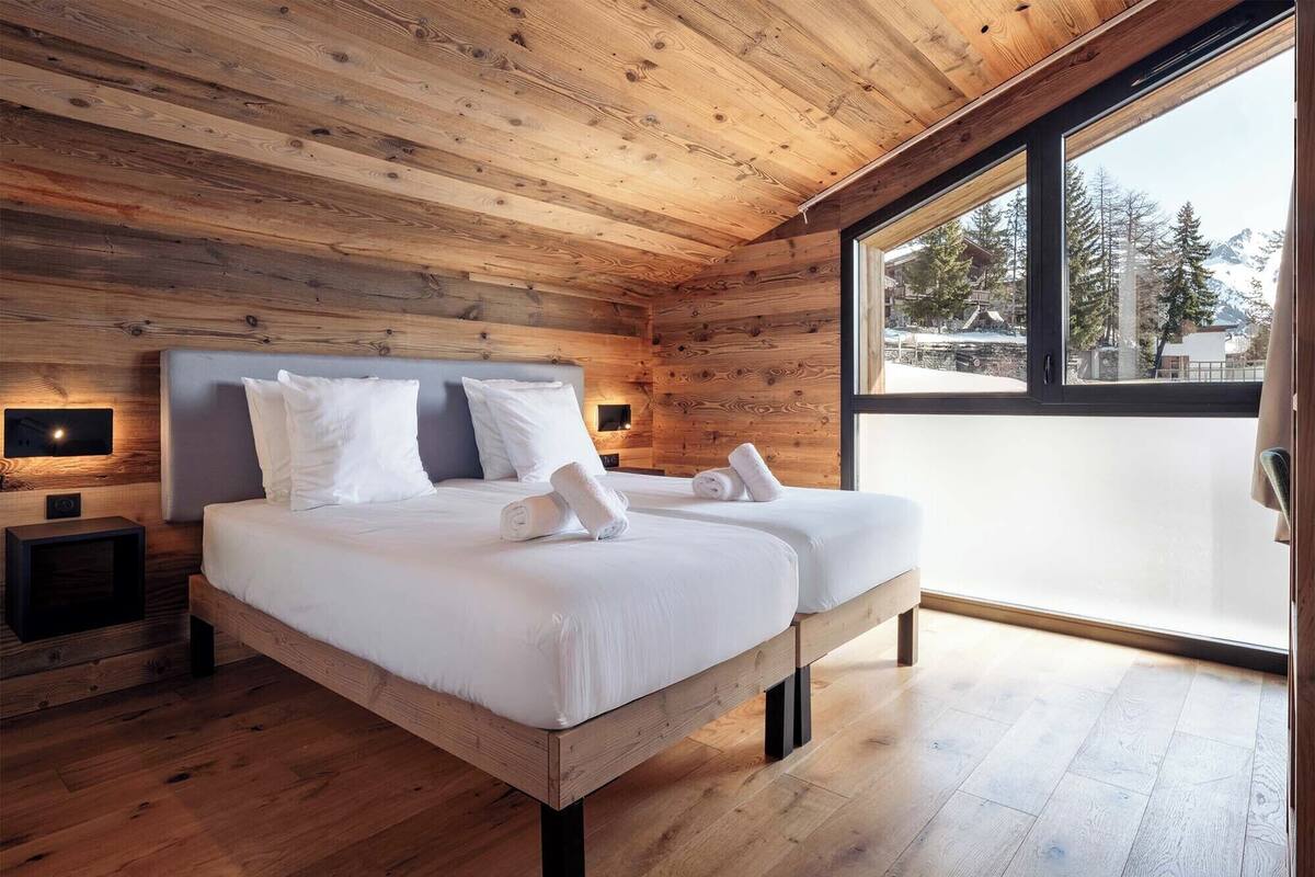 Ein schlicht eingerichtetes Schlafzimmer im Chalet-Stil mit zwei Betten, einem großen Fenster und Blick ins Freie.