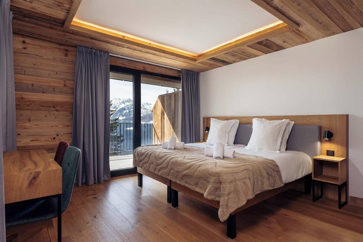 Ein modernes Hotelzimmer mit Holzboden, einem großen Bett mit weißer Bettwäsche, einem Schreibtisch mit Stuhl und Blick auf Berge durch das Fenster.
