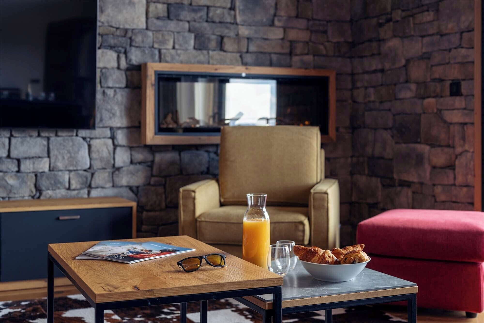A cozy living room with a stone wall, a comfortable armchair, a wooden coffee table with a magazine, a pair of glasses, a glass of orange juice, and a bowl of croissants.