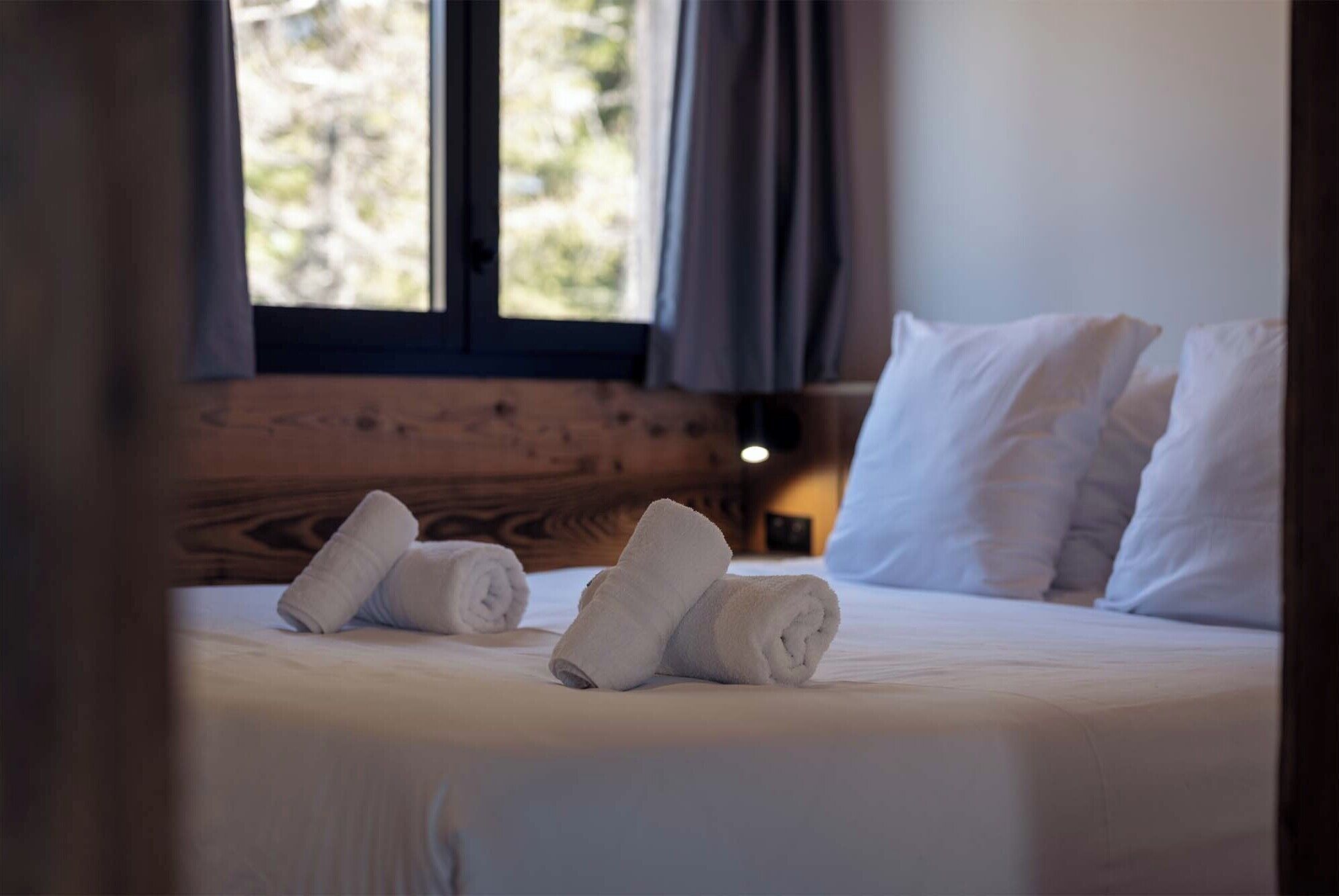 Una cama bien tendida con sábanas y almohadas blancas, un cabecero de madera y una ventana con cortinas.