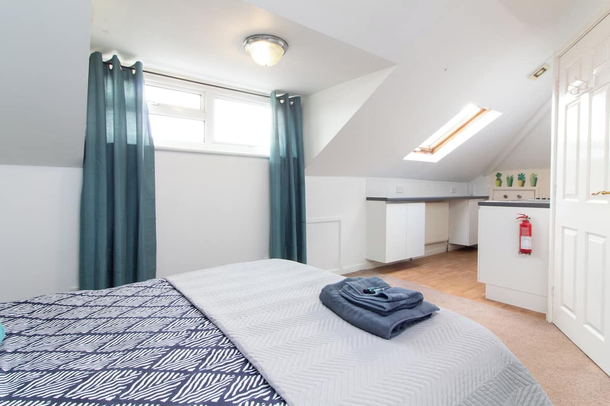 A bedroom with a bed, a window with curtains, a kitchenette, and a ceiling light.