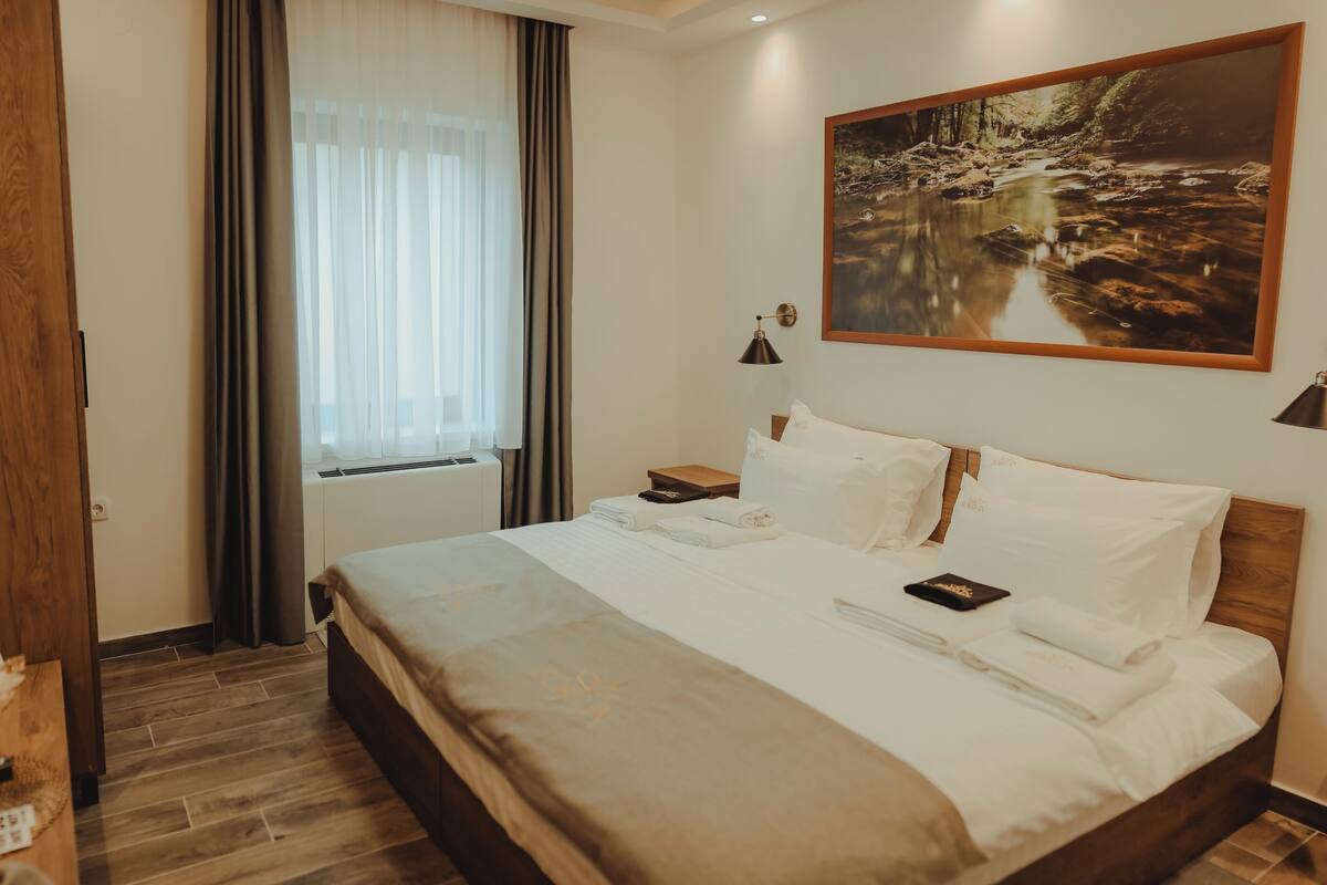 A neatly made bed with white linens, a wooden headboard, and a painting of a forest stream on the wall.