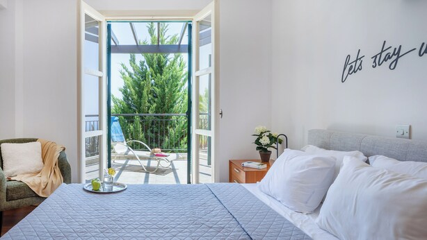 A hotel room with a bed, a chair, a small table with a tray, and a balcony with a view of trees. - J1 Holiday House Adjacent to Aphrodite Hills - Sea View (Kouklia)