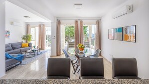 A modern living room with a sofa, coffee table, and chairs. There is a dining table with a fruit bowl. The room has large windows with curtains, an air conditioning unit, and artwork on the wall. - J1 Holiday House Adjacent to Aphrodite Hills - Sea View (Kouklia)