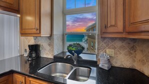 A kitchen with a double sink, wooden cabinets, a coffee maker, and a window with a view of the ocean and buildings.