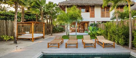 A poolside area with lounge chairs, a wooden deck, and a thatched-roof building.