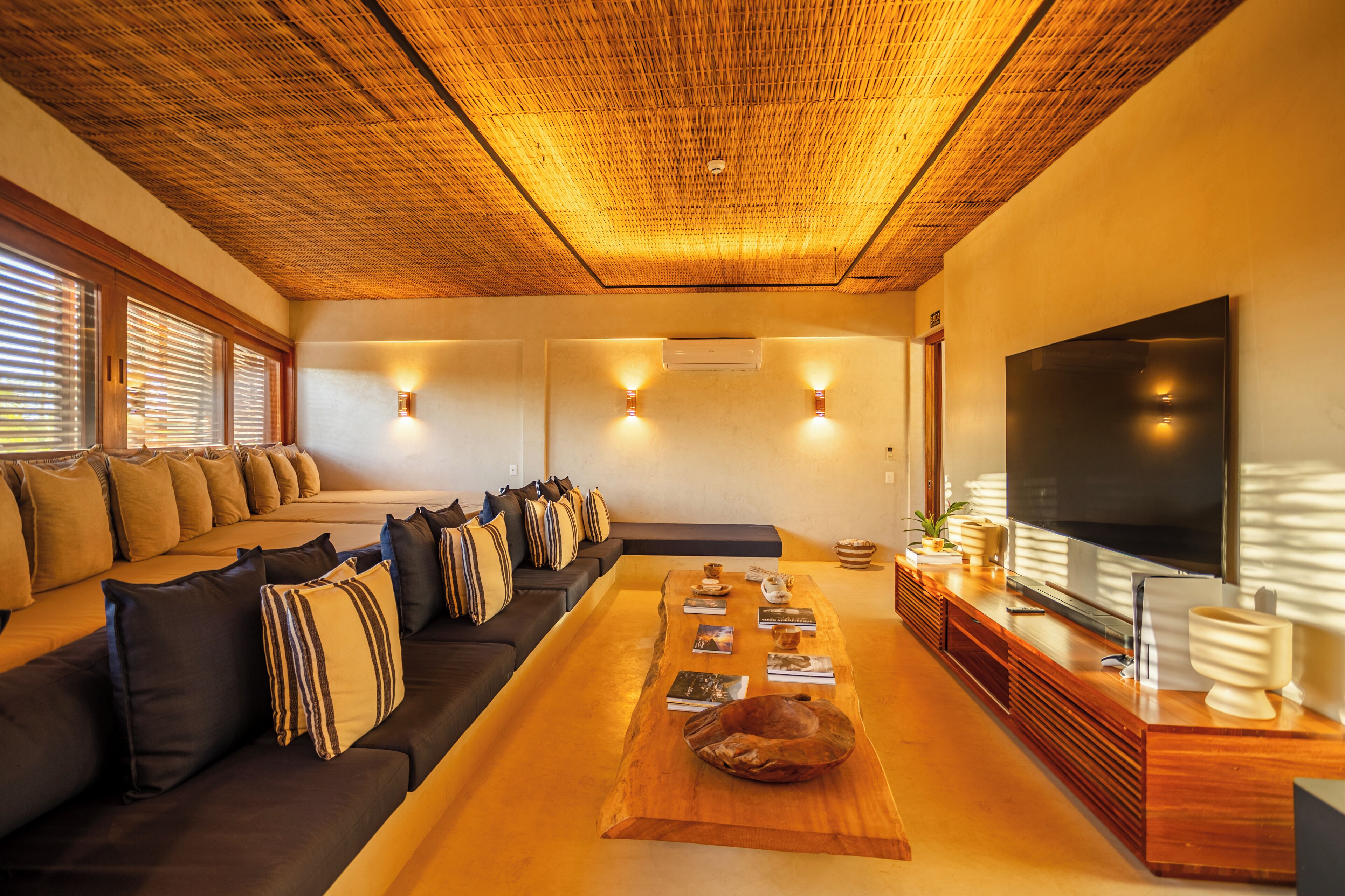 Uma sala de estar moderna com um sofá de madeira comprido, uma mesa de centro com livros, uma TV de tela plana e luminárias fixadas na parede.