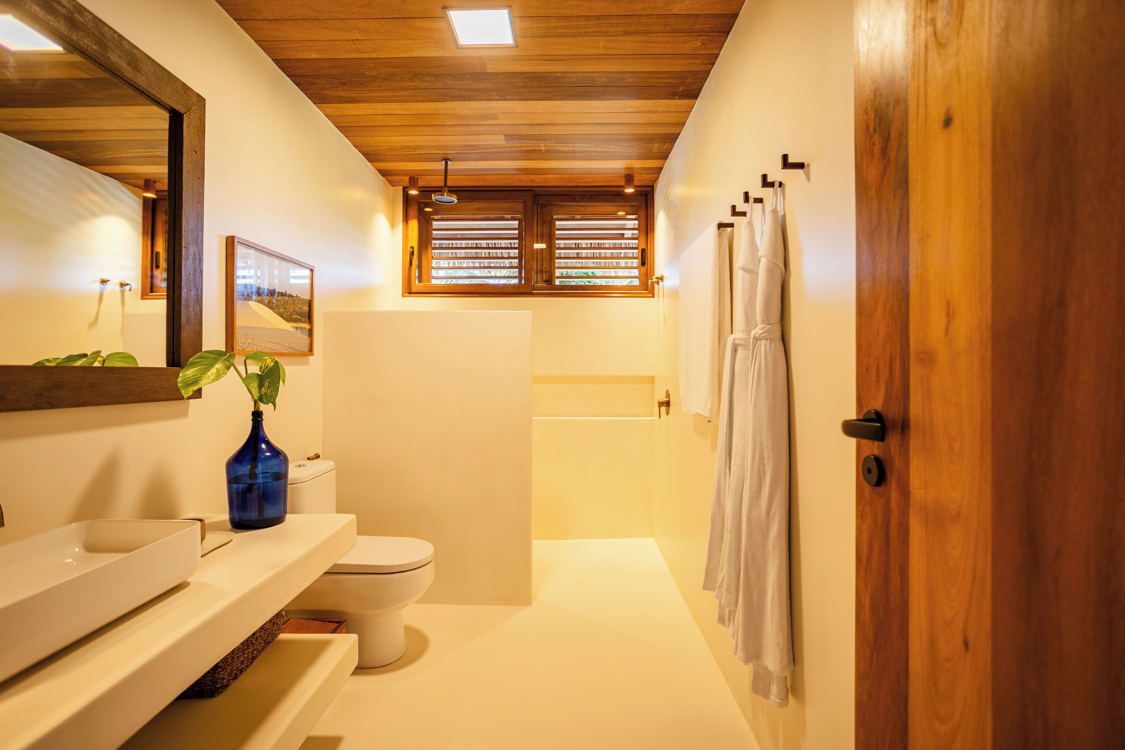 A modern bathroom with a wooden ceiling, a white sink, a toilet, a bathtub, and a window with blinds.