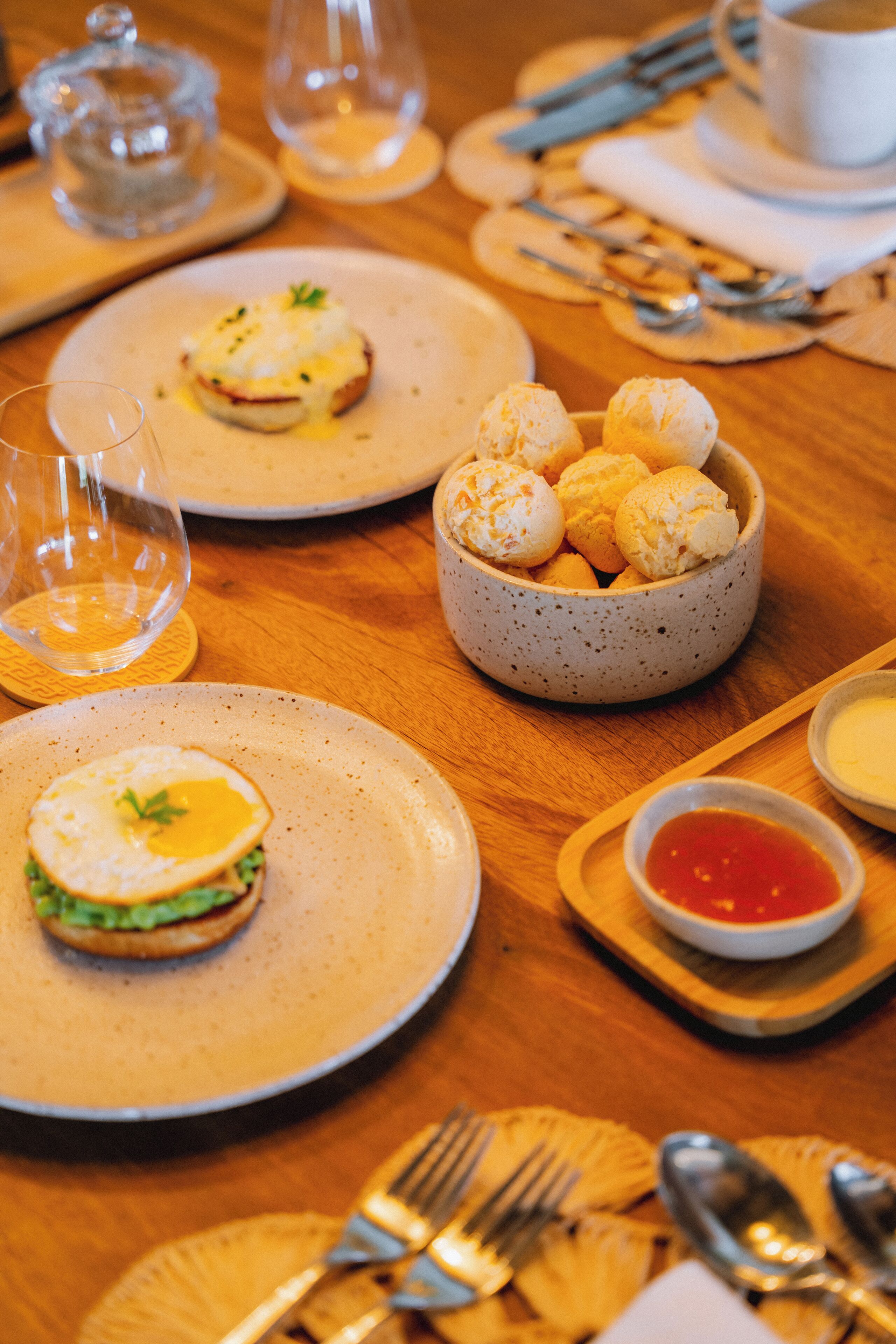 Une table à manger dressée avec des assiettes remplies de nourriture, notamment un œuf poché et un sandwich, un bol de petits pains et un petit bol de sauce.