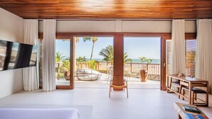 A hotel room with a wooden ceiling, a large bed, a TV mounted on the wall, a balcony with a view of the ocean, and a small desk with chairs. - Casa Siará - Preá (Cruz)