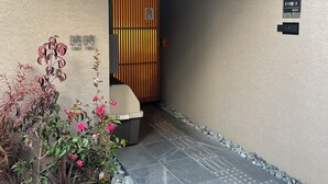 Une entrée moderne avec une porte à lattes en bois, un petit espace extérieur aménagé en coin salon et un chemin de pierre menant à l’entrée.