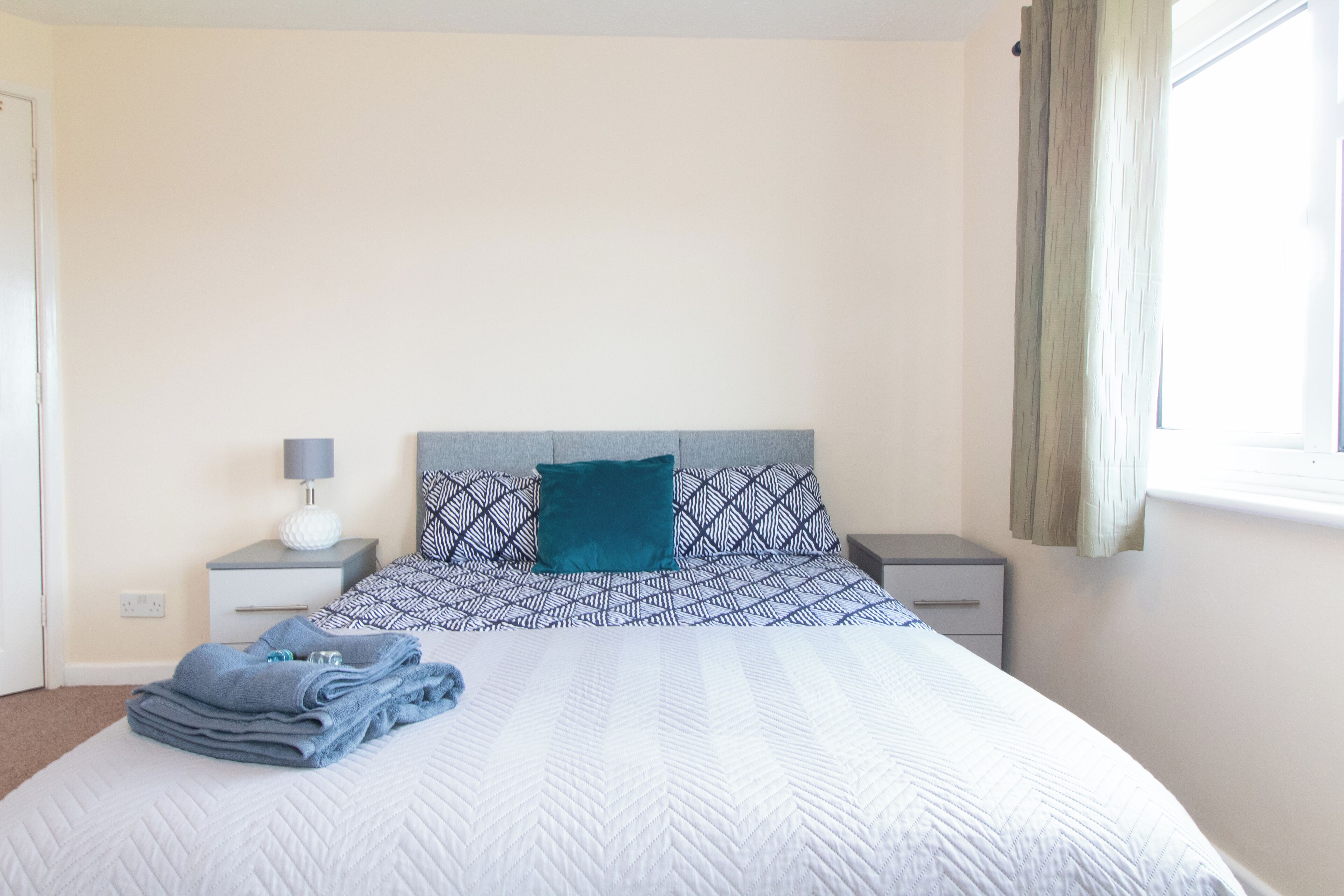 A neatly made bed with a patterned bedspread, a teal pillow, and folded towels on a bedside table with a lamp.