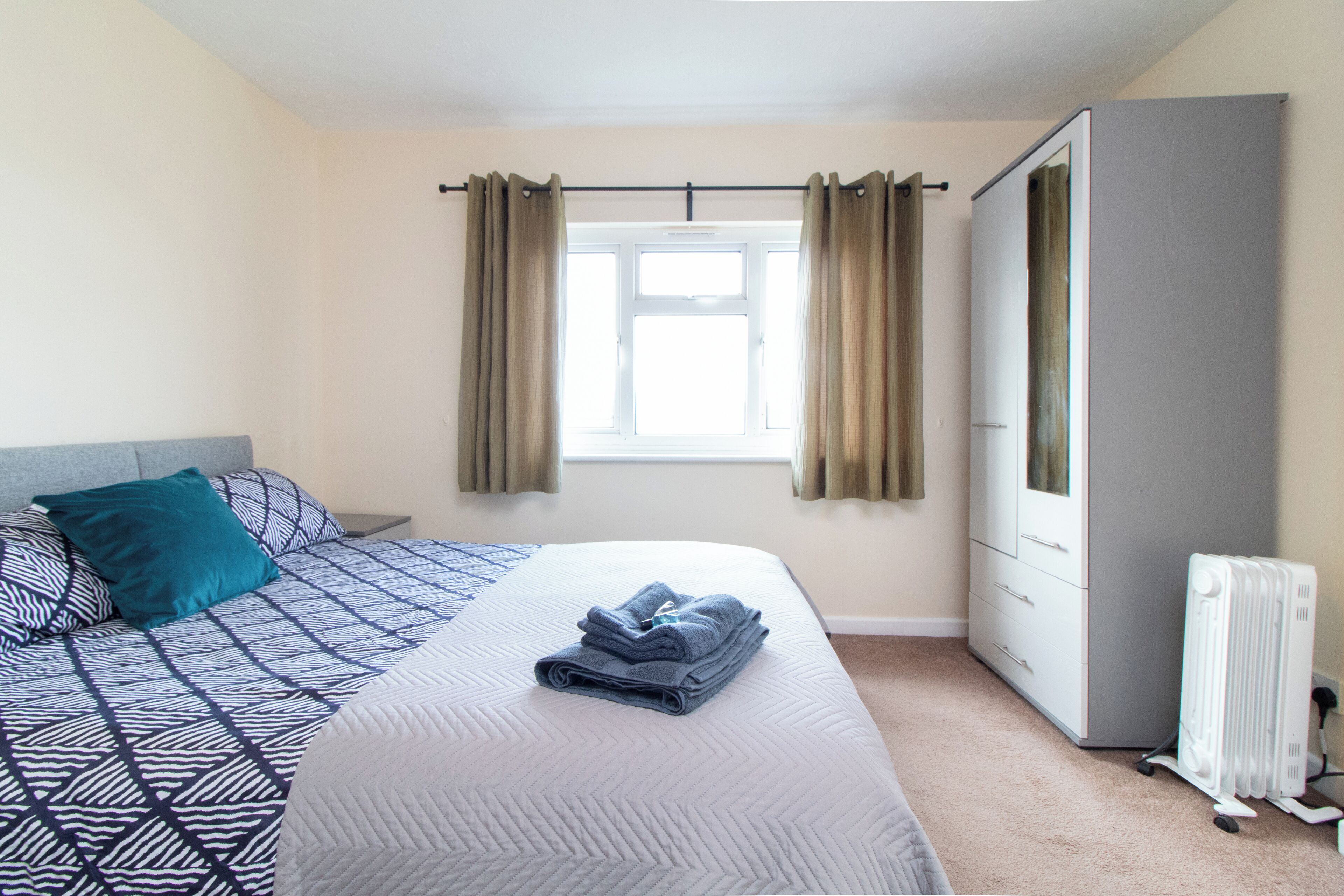 Un dormitorio con cama, radiador, armario y ventana con cortinas.