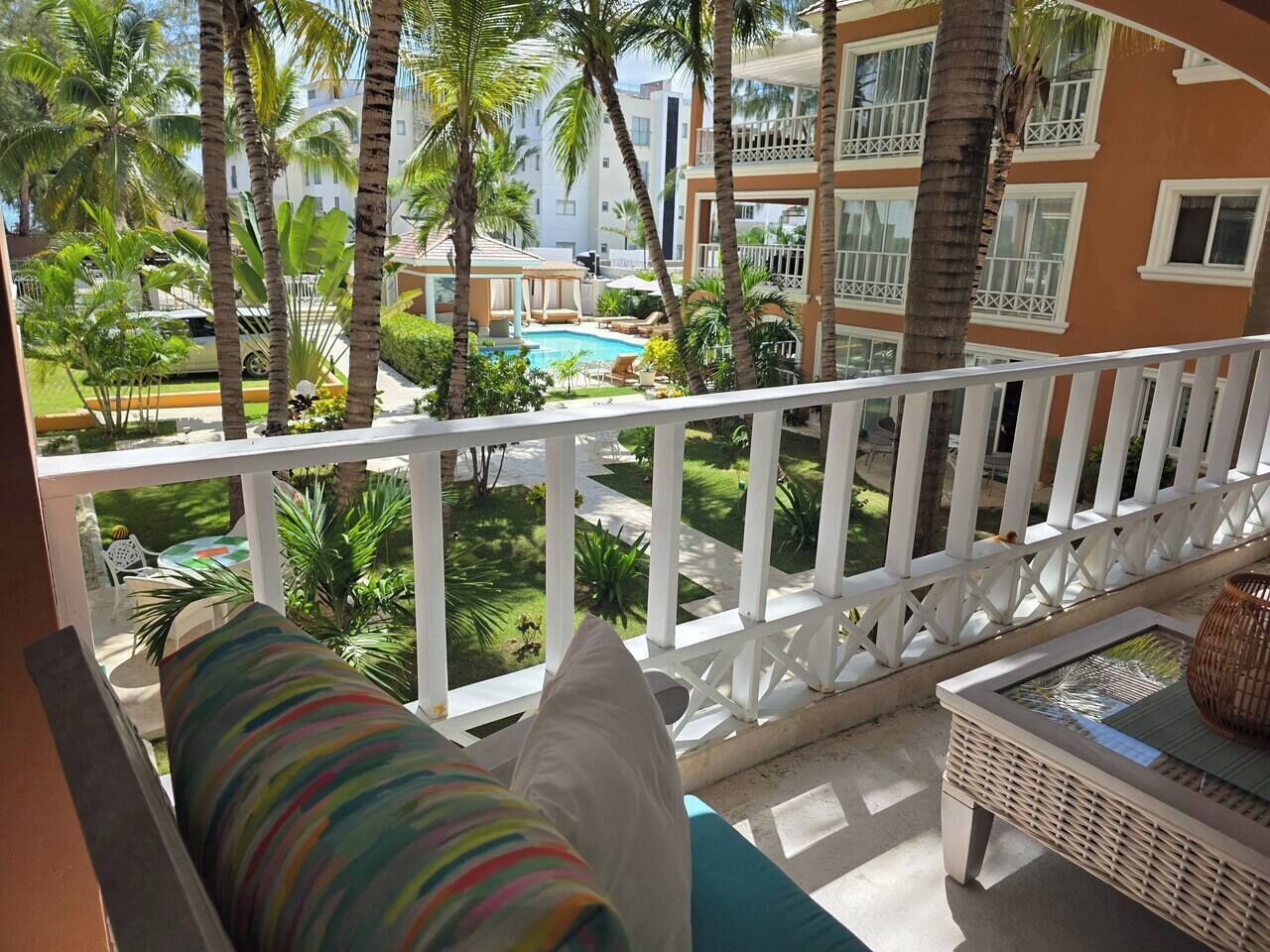A balcony with a view of a swimming pool, palm trees, and a multi-story building with balconies.