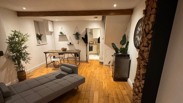 A spacious living area with a wooden floor, a grey sofa, a dining table with chairs, and a kitchen visible through an open doorway.