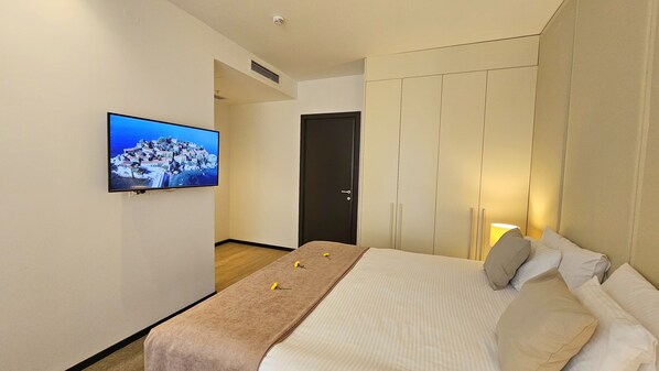Habitación de hotel con cama, televisor montado en la pared, lámpara de noche y puerta que da a otra habitación.