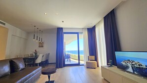 A modern living room with a sofa, a coffee table, a TV, and a balcony with a view of the sea. - Royalis Suites Rafailovici - Beachfront Sea View Apartments (Becici)