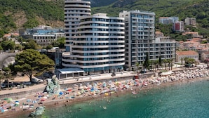 A coastal hotel with multiple floors, a beach with umbrellas, and a mountainous backdrop. - Royalis Suites Rafailovici - Beachfront Sea View Apartments (Becici)
