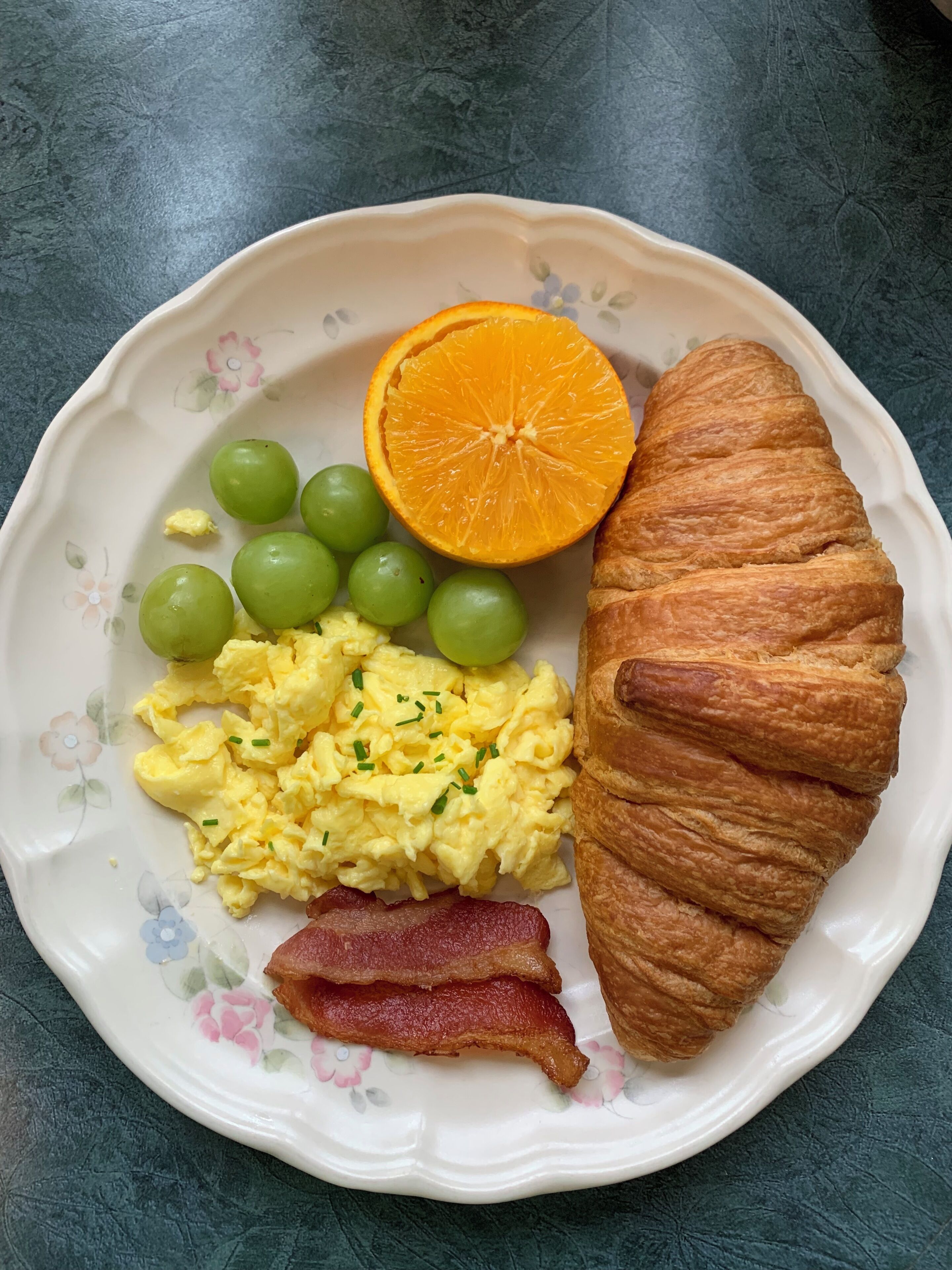 A plate with scrambled eggs, bacon, green grapes, an orange slice, and a croissant.