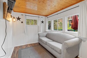Una habitación con techo de madera, un sofá gris, una ventana con vistas a árboles y una lámpara fijada a la pared.