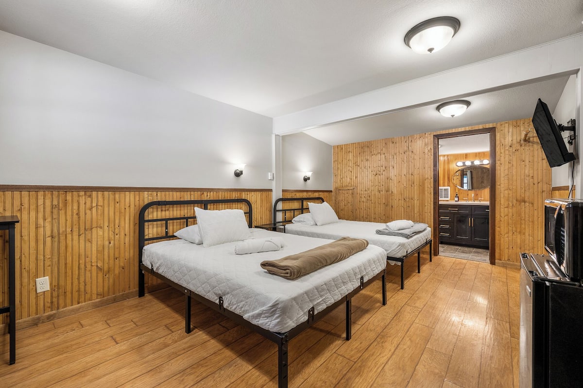 A room with two beds, wooden walls, a kitchen area, and a TV.
