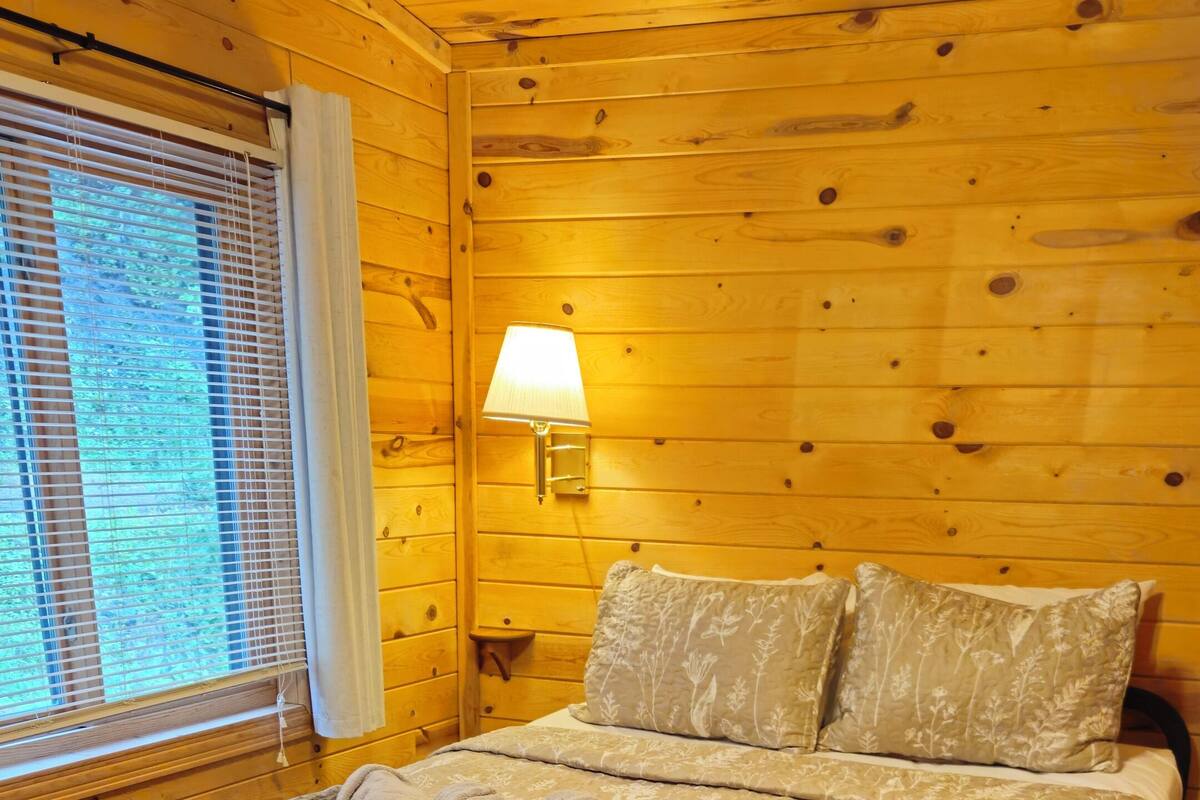 A wooden cabin bedroom with a bed, pillows, a lamp, and a window with blinds.