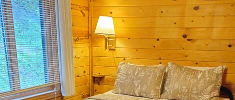 A wooden cabin bedroom with a bed, pillows, a lamp, and a window with blinds.