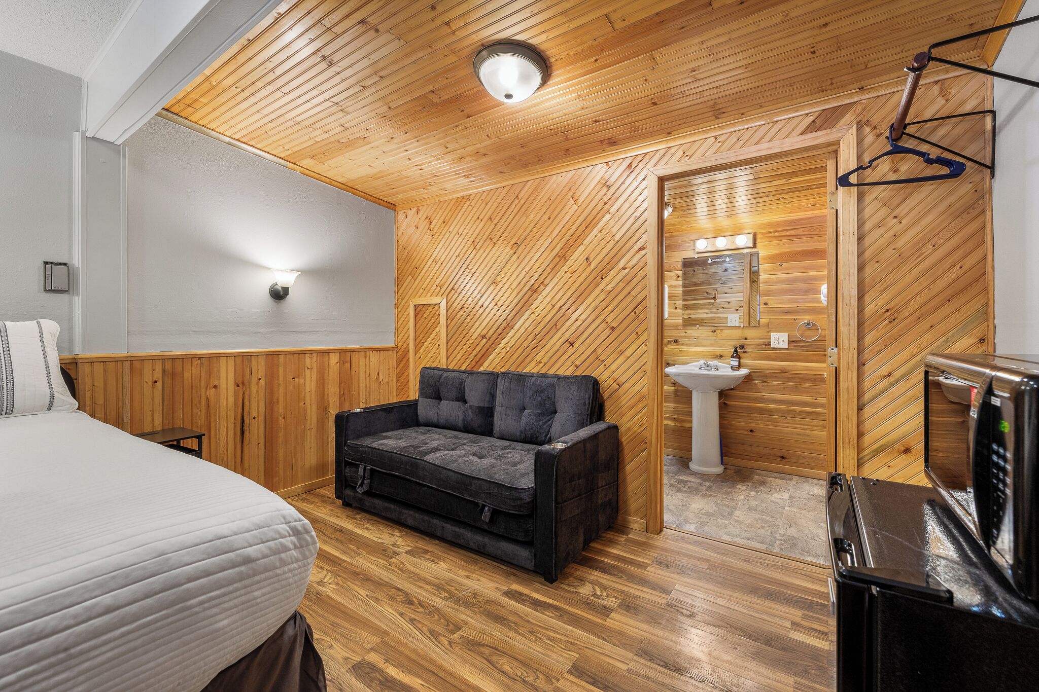 A compact room with wooden walls and ceiling, a bed, a dark sofa, a small table, a sink, and a microwave.