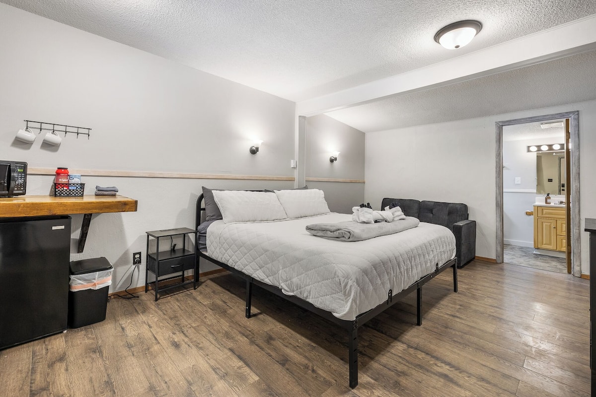 A bedroom with a bed, a desk, a trash bin, and a mirror.