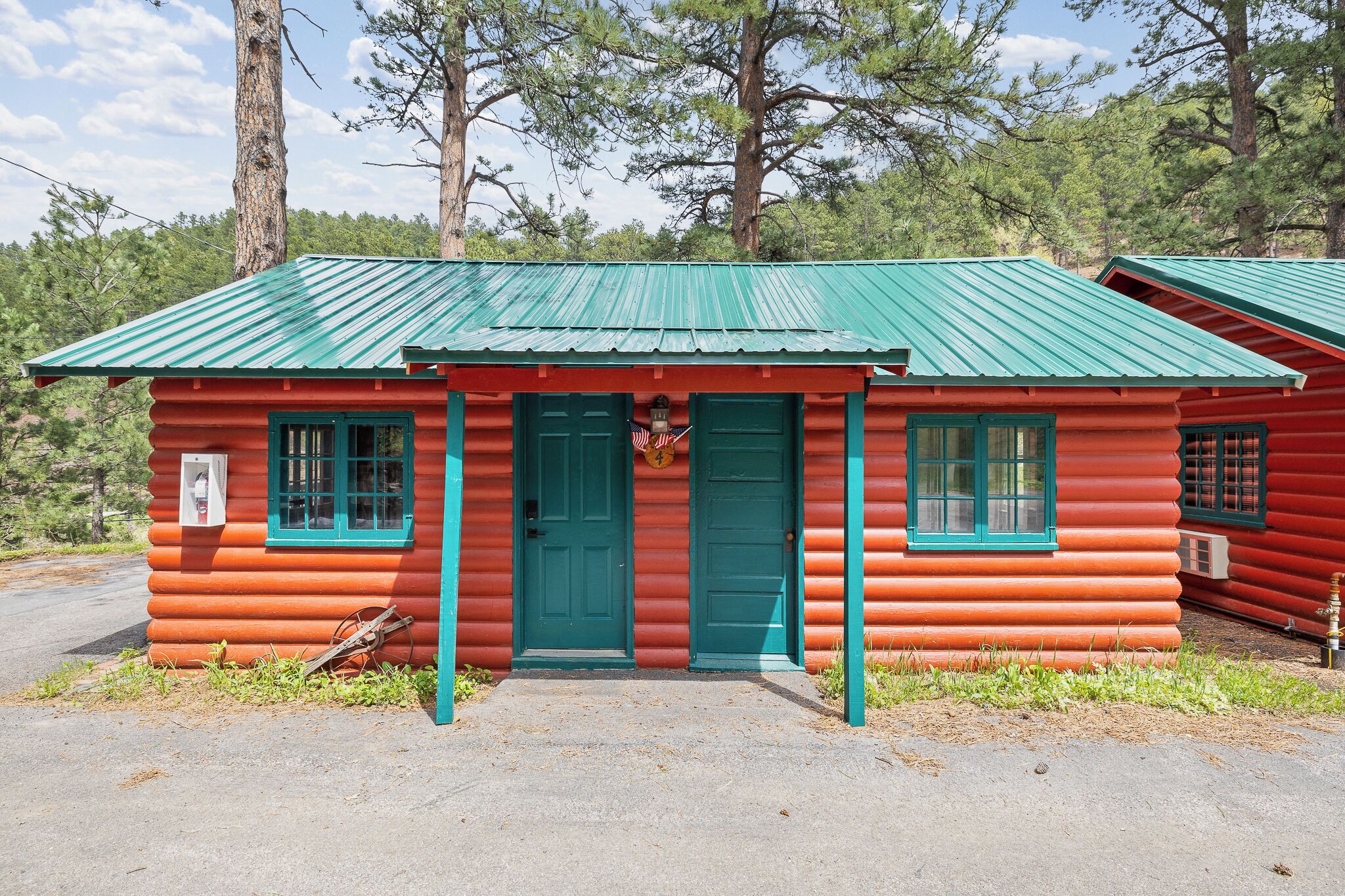 A log cabin with a green metal roof and a red exterior, featuring two green doors and windows, and a small American flag.