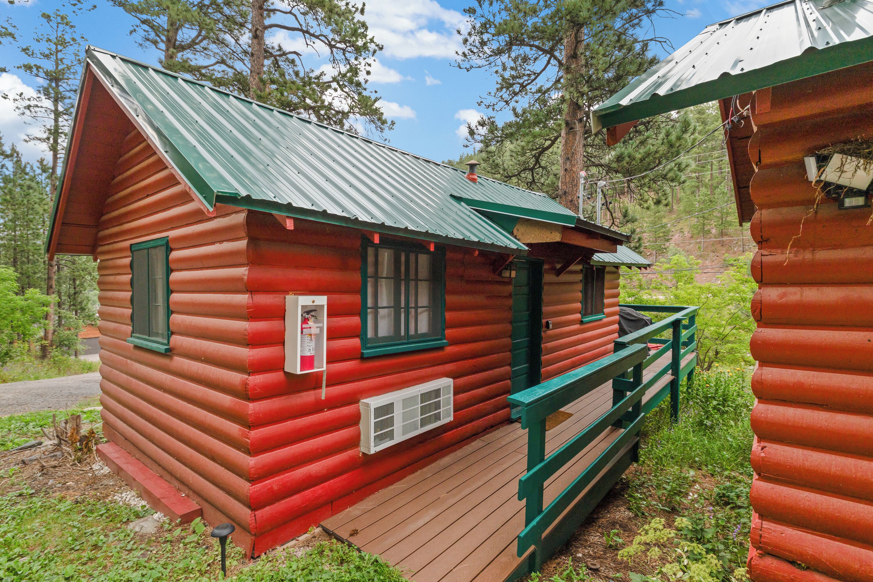 A red log cabin with a green roof, a wooden deck, and a small porch.