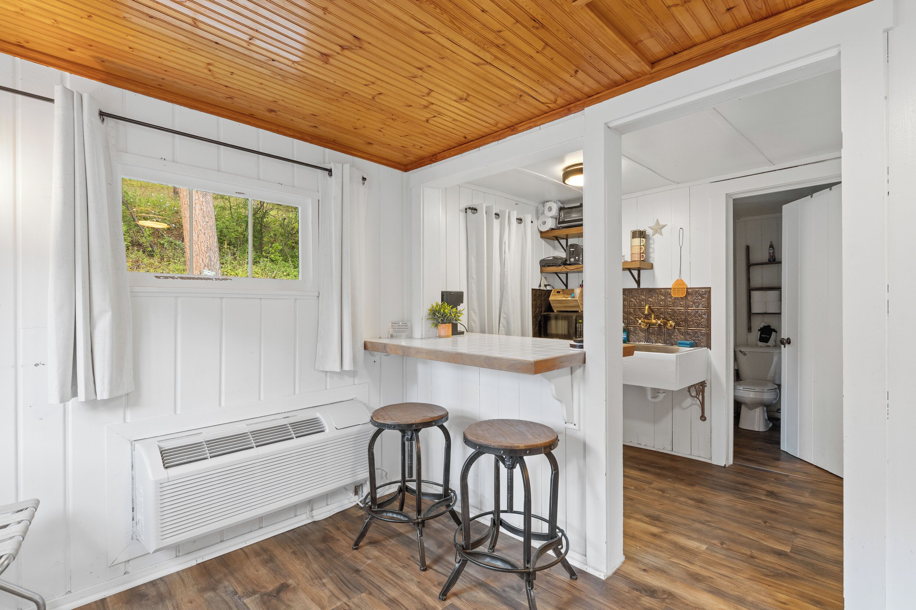 A kitchen with a wooden ceiling, white walls, and a window. There are two stools and a sink with a wooden countertop.