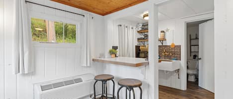 Eine Küche mit Holzboden, weißen Wänden und einem Fenster. Zwei Barhocker und eine Spüle mit einer Holzarbeitsplatte sind vorhanden.