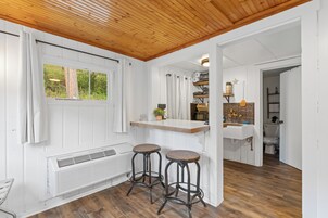 Cocina con techo de madera, paredes blancas y ventana. Hay dos taburetes y un lavamanos con encimera de madera.