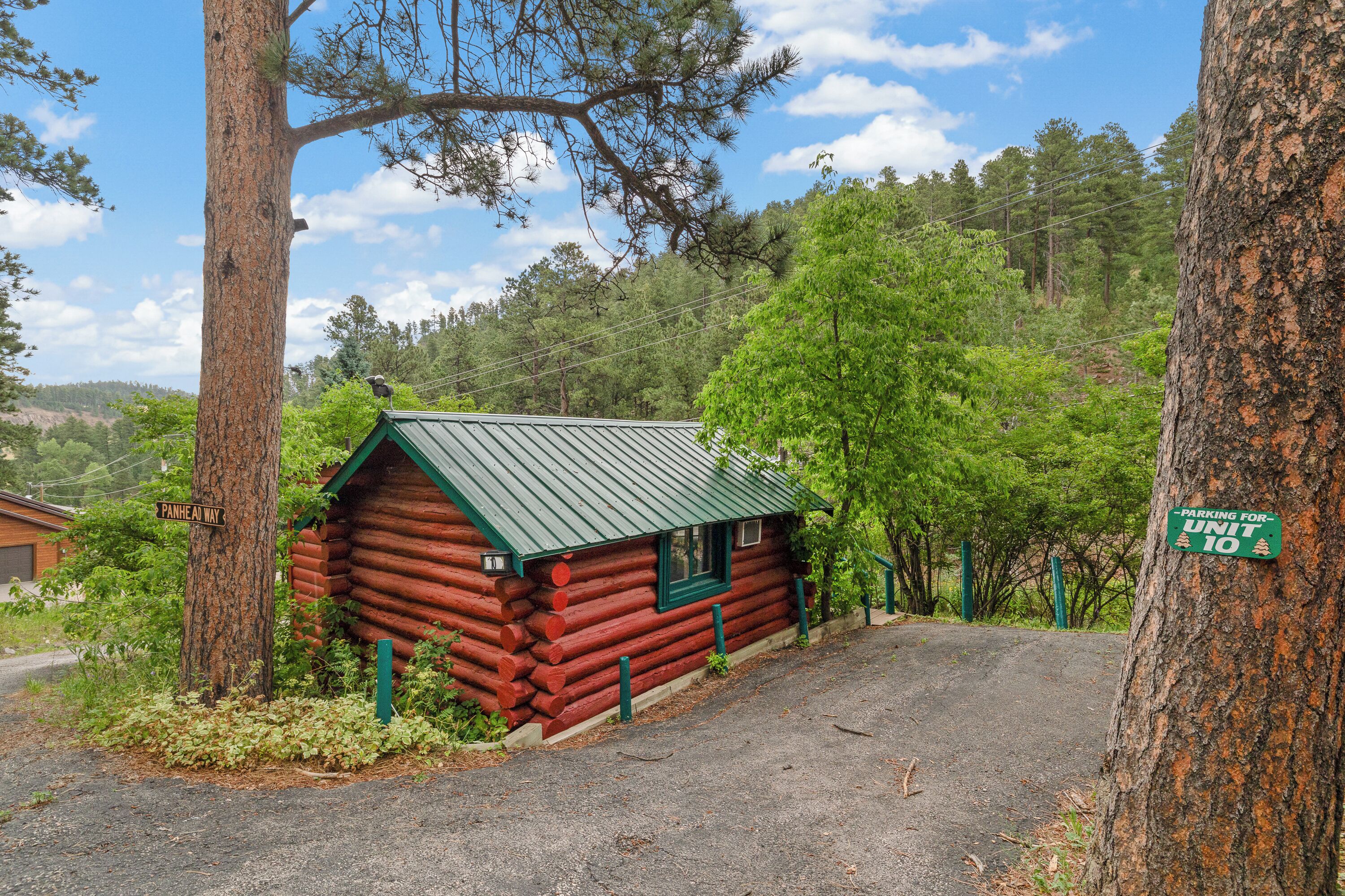 A small log cabin with a green roof, surrounded by trees and a gravel driveway.