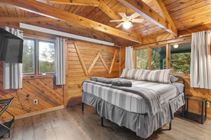 Un dormitorio de cabaña de madera con una cama, un ventilador de techo y ventanas amplias.