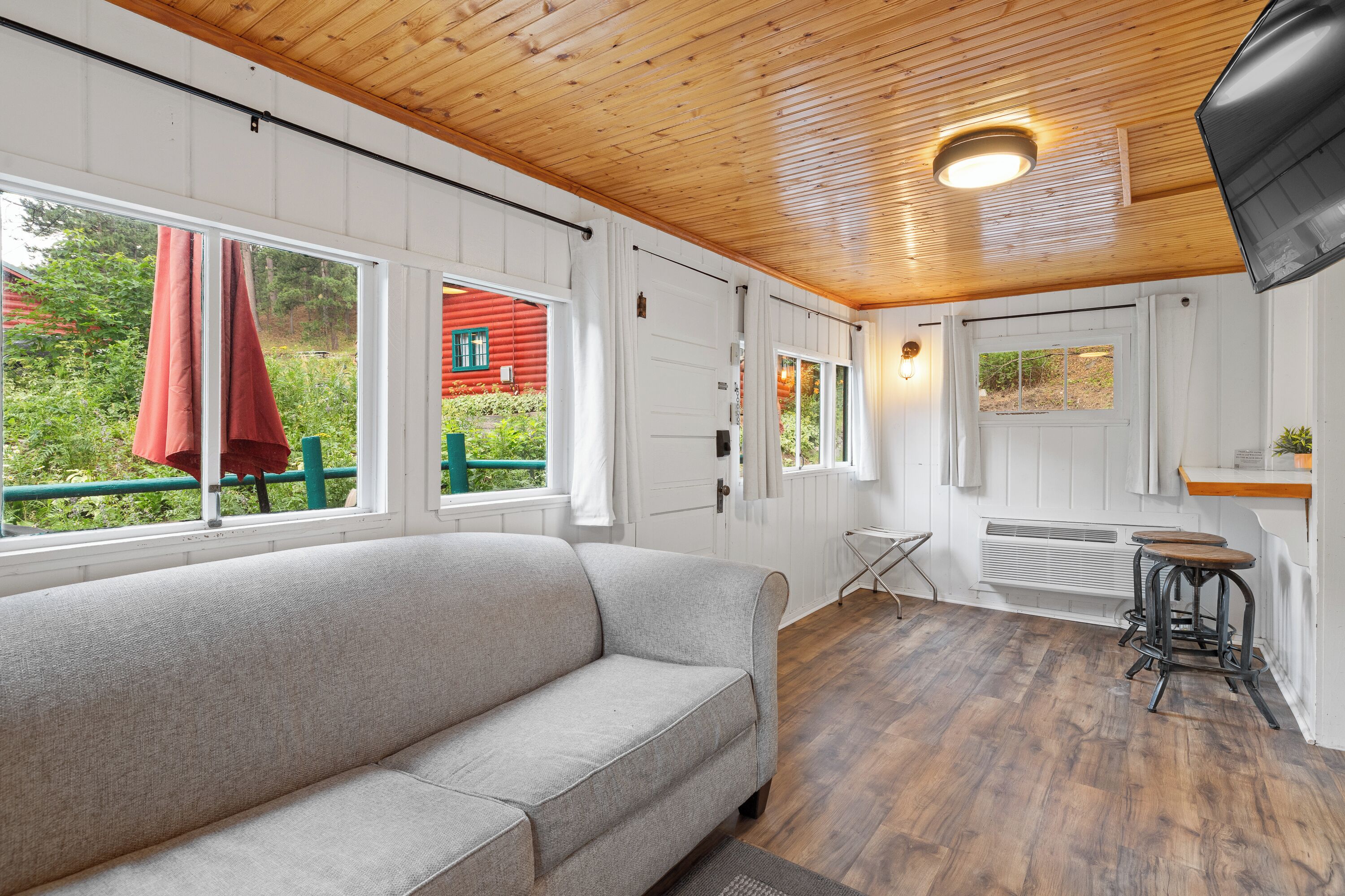 A cozy room with a sofa, wooden flooring, a window with a red curtain, and a view of greenery outside.