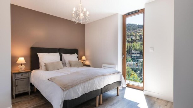 A bedroom with a large bed, bedside lamps, a chandelier, and a view of a balcony with a cityscape. - Les Charmettes (Brides-les-Bains)