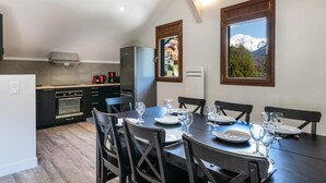 A modern kitchen with dark cabinetry, a built-in oven, and a stainless steel refrigerator. There is a dining table set with plates and glasses, and a window with a mountain view. - Les Charmettes (Brides-les-Bains)