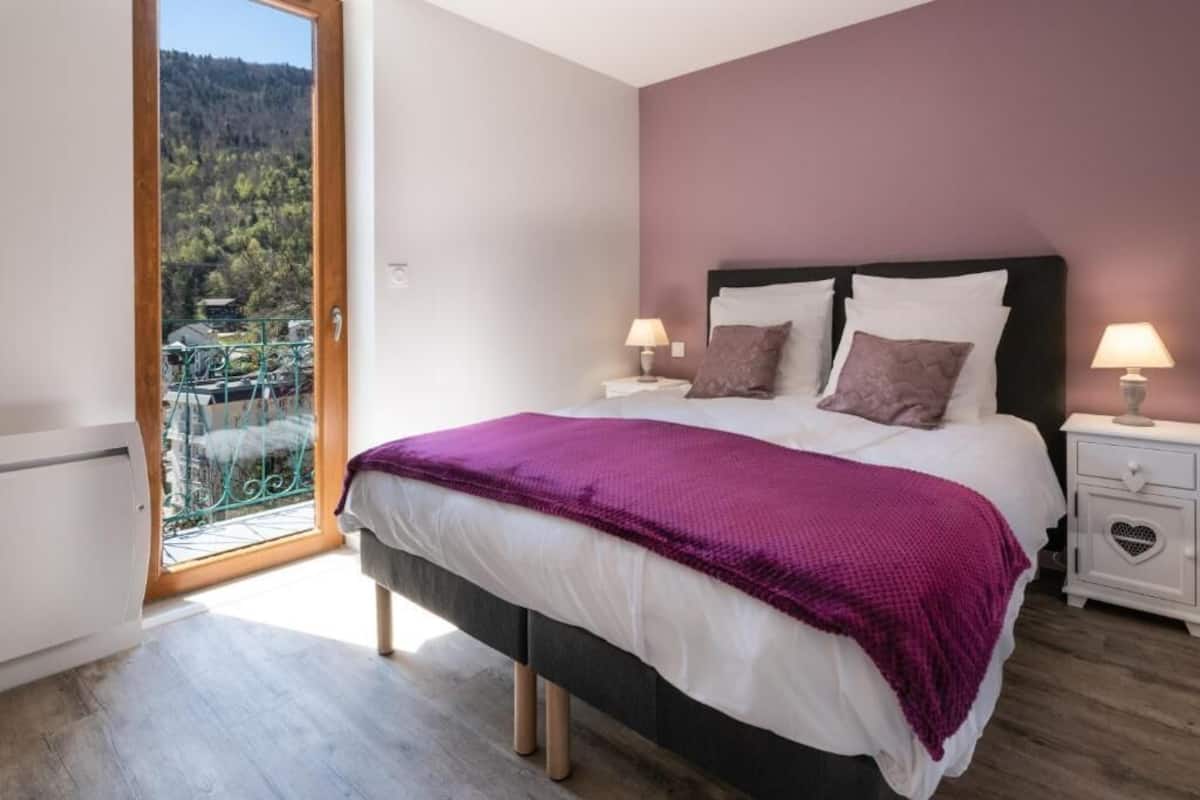 Un dormitorio con una cama, dos mesitas de noche, una ventana que ofrece vistas a un balcón y árboles, y una puerta.