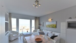 A modern, well-lit living area with a dining table, white sofas, and a large window offering a view of the outdoors. - Studio in Duinbergen by the Sailing Club (Knokke-Heist)