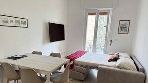 A compact room with a bed, a sofa, a dining table with chairs, a TV, and a window with a view of buildings.