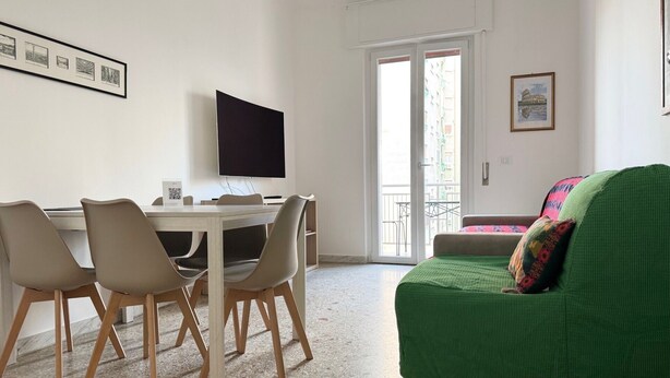 A room with a dining table, chairs, a TV, a green sofa, and a balcony with a view of buildings.