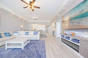 A modern living room with a white sofa, blue accent pillows, a coffee table, and a large TV screen displaying a beach scene.